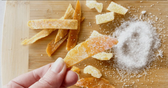 What makes sour candy sour A hand holding candied orange peel over a pile of citric acid and sugar.
